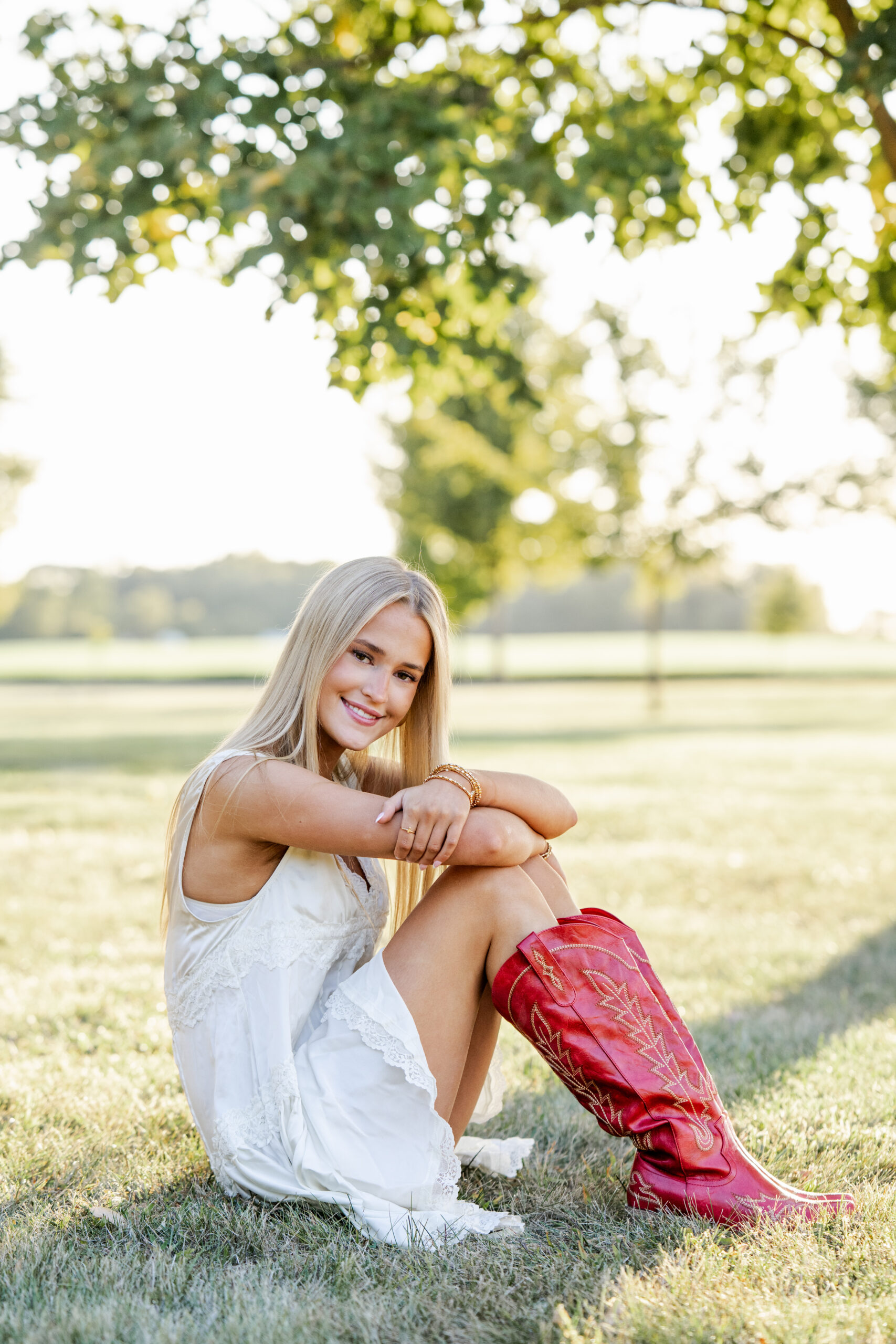 Senior girl in red cowboy boots sits in the grass during Sheridan, Indiana senior photo session.